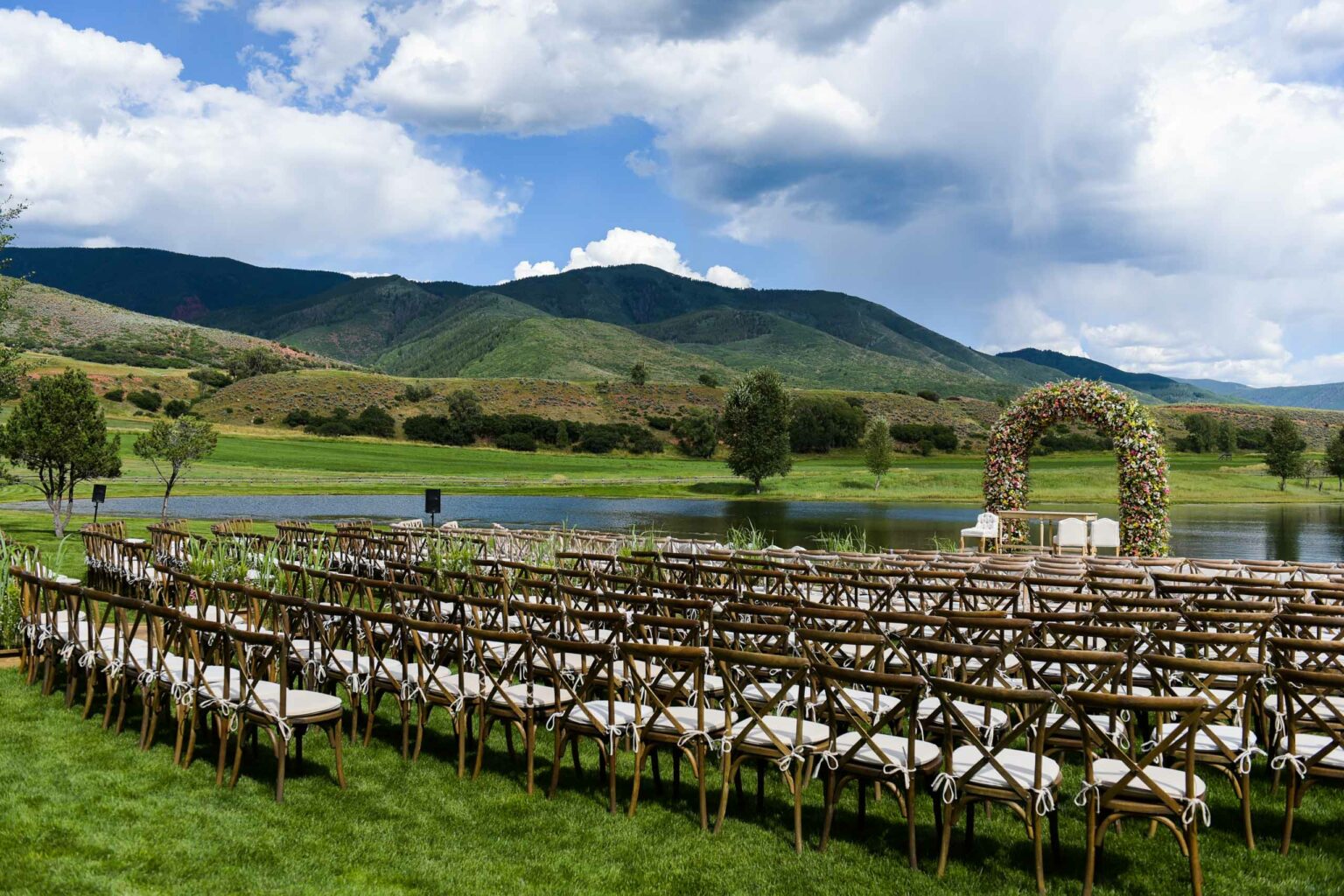 chairs set out for a wedding ceremony outside next to a lake with a floral arch.