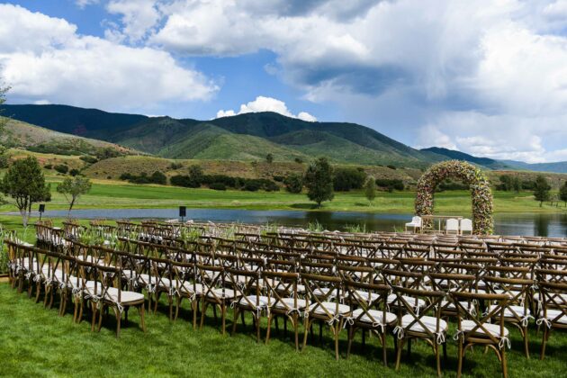 Outdoor wedding venue set up by professional event planners with rows of chairs and a floral arch near a tranquil lake with mountains in the background.