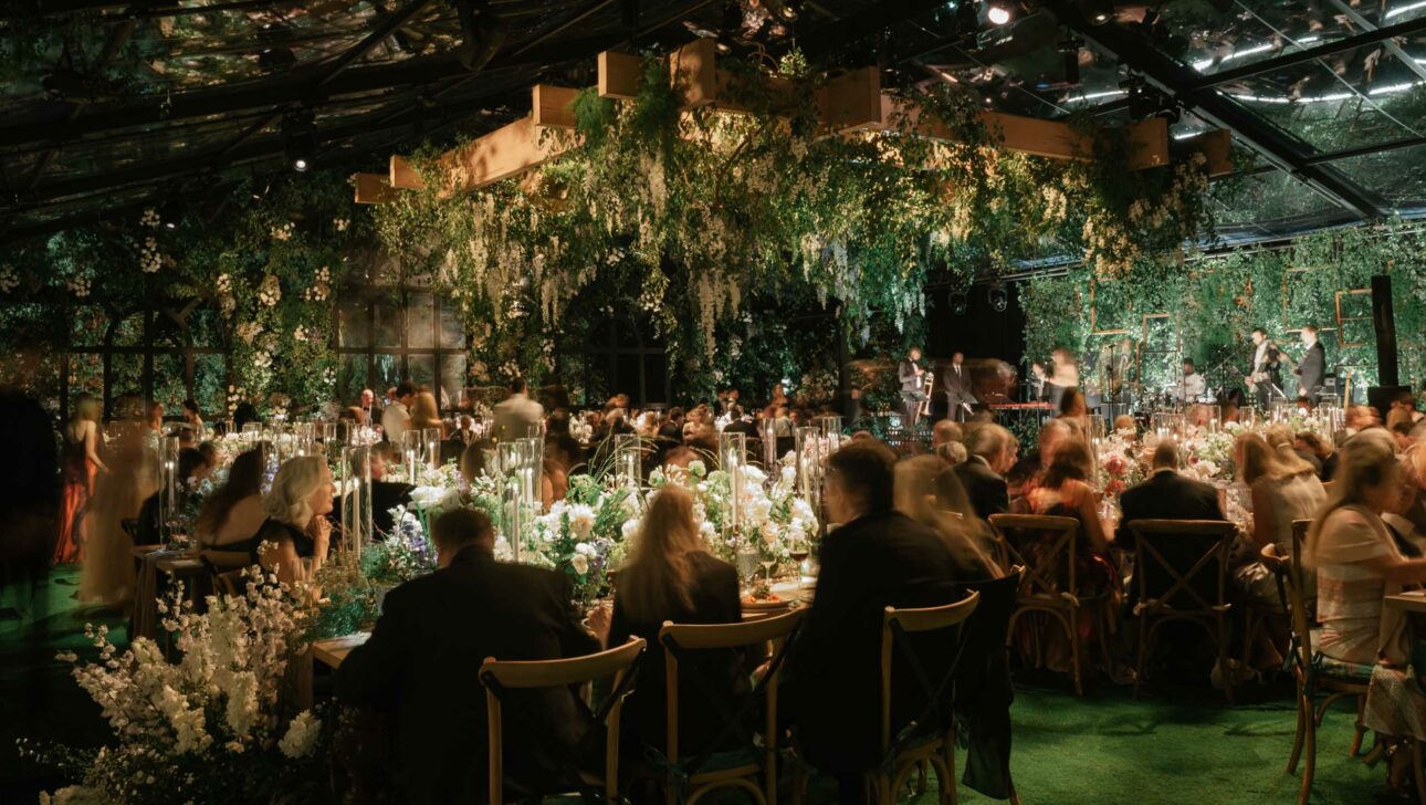 An elegant event planned by a top event planning company, with guests seated at tables adorned with white floral arrangements, set in one of their exclusive event venue spaces with lush greenery overhead, creating an enchant