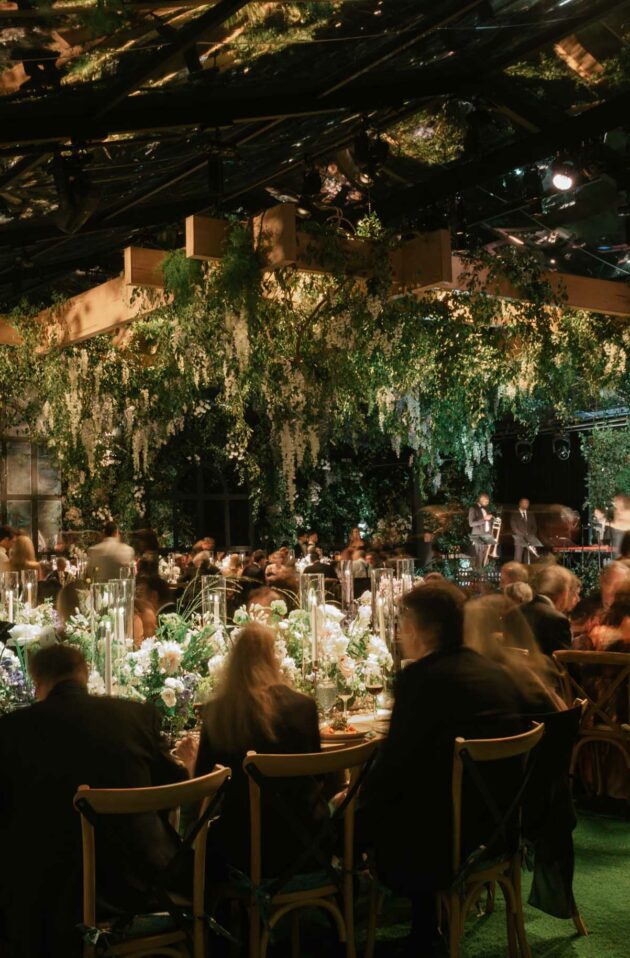 An elegant event planned by a top event planning company, with guests seated at tables adorned with white floral arrangements, set in one of their exclusive event venue spaces with lush greenery overhead, creating an enchant