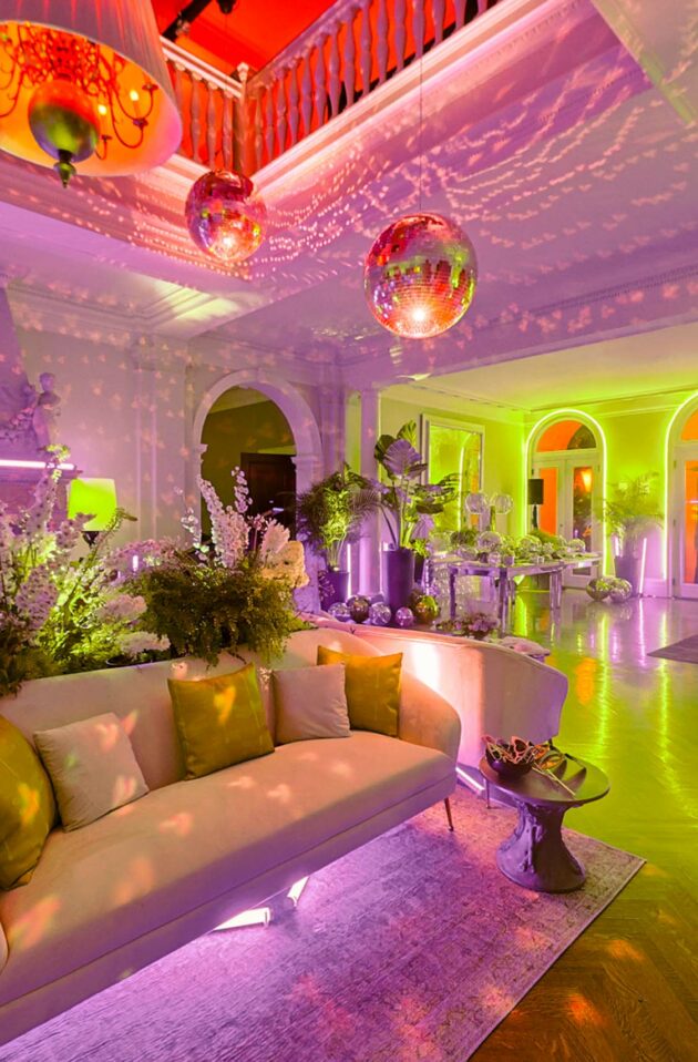 An elaborately decorated room lit with pink and green lights, managed by an event planning company, featuring a sofa, floral arrangements, and reflective disco balls.