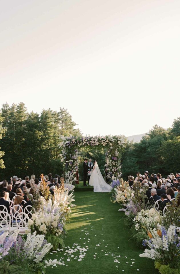 Outdoor wedding ceremony organized by event planners, with guests seated on either side of a green aisle leading to a floral arch, framed by trees under a clear sky.