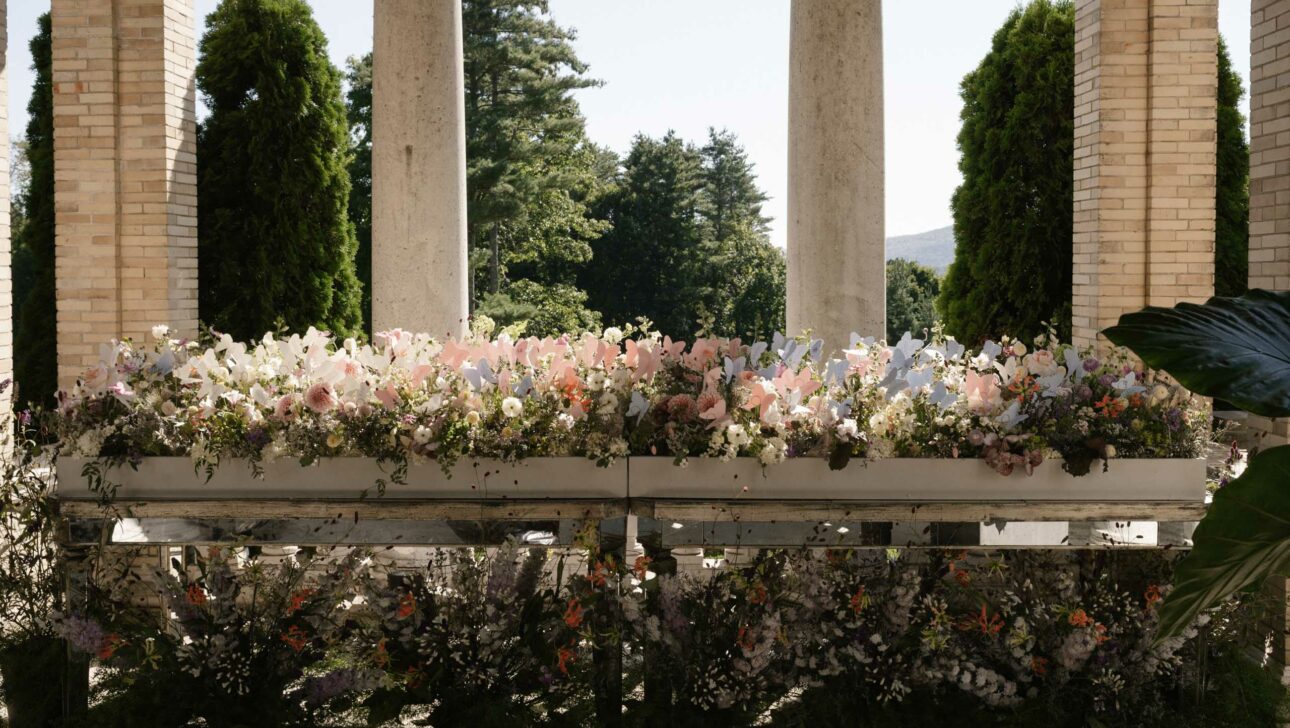 An array of floral arrangements displayed between classical columns at an event venue space, with a backdrop of trees and a clear sky.