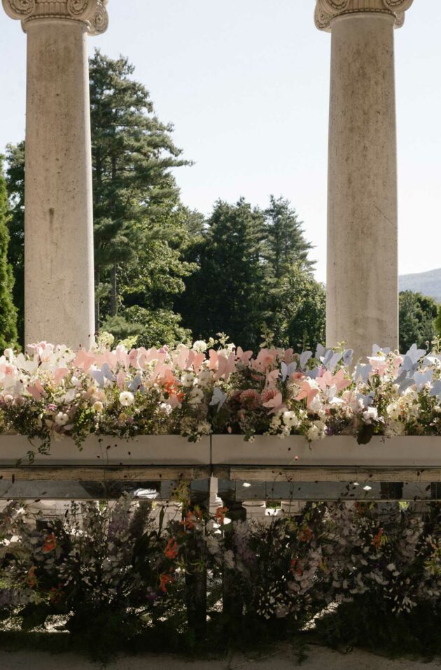 An array of floral arrangements displayed between classical columns at an event venue space, with a backdrop of trees and a clear sky.