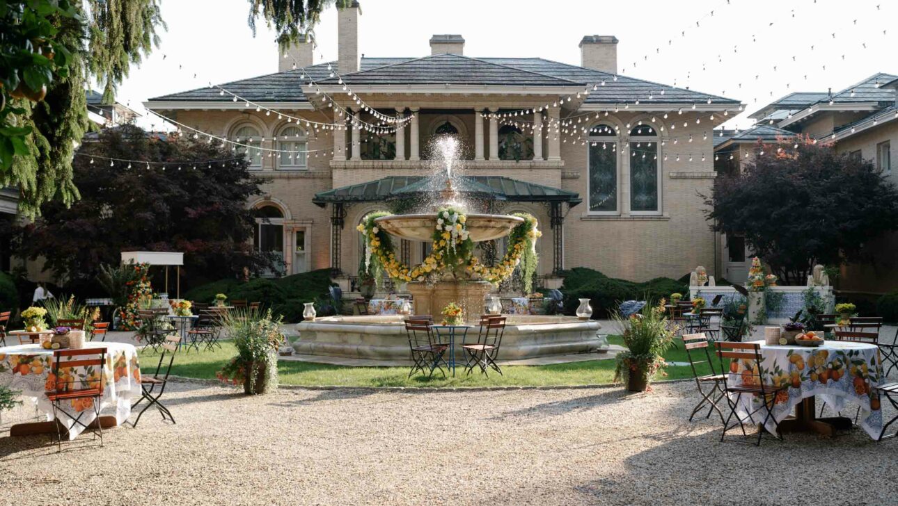 Outdoor event setup by an event planning company, with tables and a fountain in front of a large house, adorned with string lights and floral decorations.