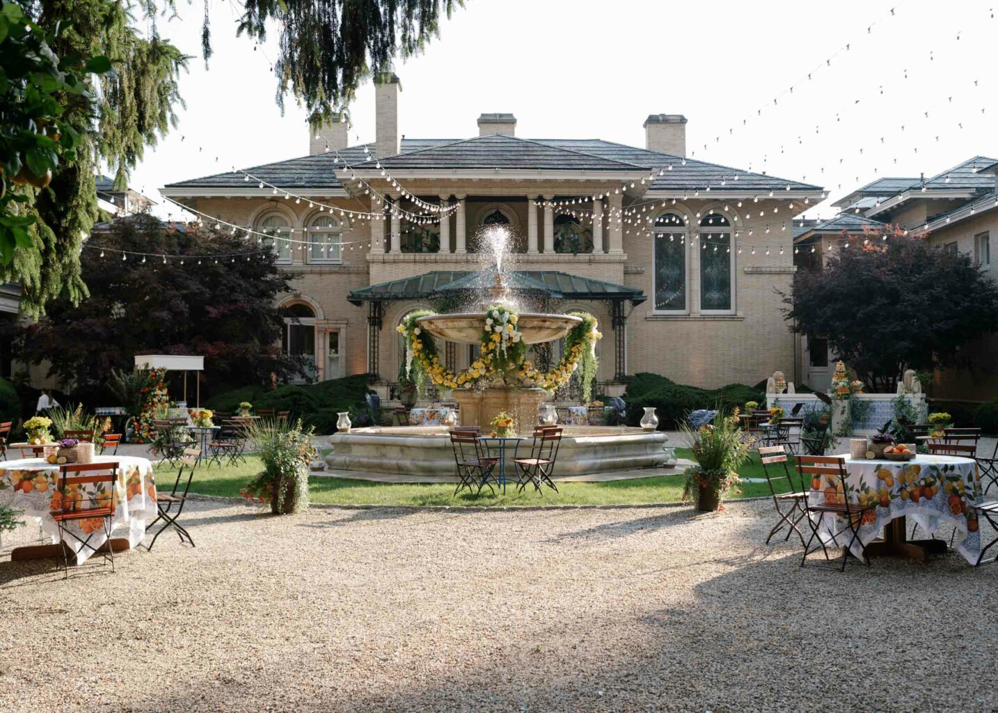 Outdoor event setup by an event planning company, with tables and a fountain in front of a large house, adorned with string lights and floral decorations.