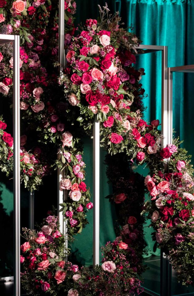 Floral arrangements adorn metal frames against a backdrop with teal drapery at the event venue space.