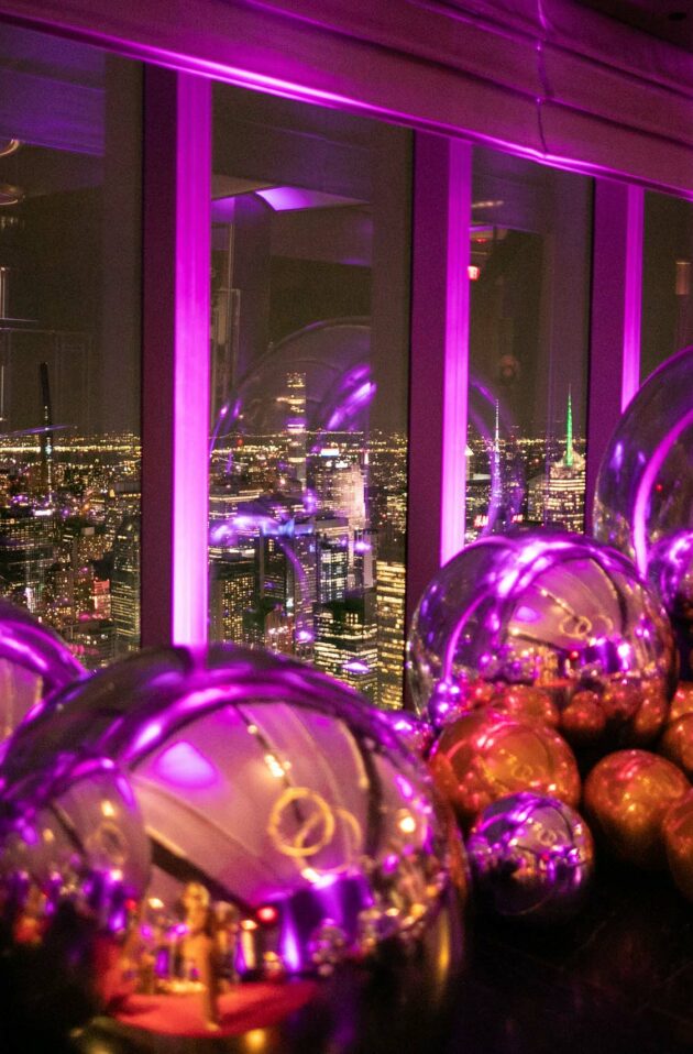 large reflective balls on the floor in a dark neon lit room looking out windows over a city skyline.