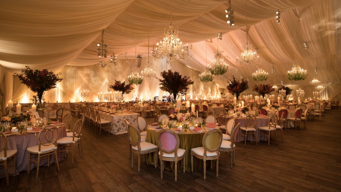 An elegantly decorated event venue space with chandeliers, draped ceiling, and tables set for an event.