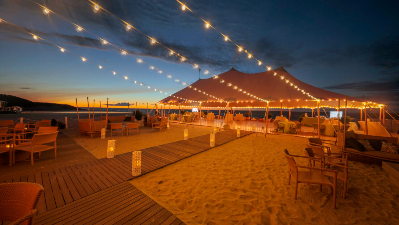 An outdoor evening event venue space lit by string lights under a large tent on a sandy beach at dusk.