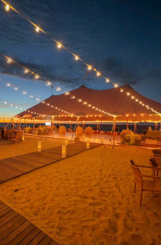 wedding reception setup under a large tent on a beach at dusk.
