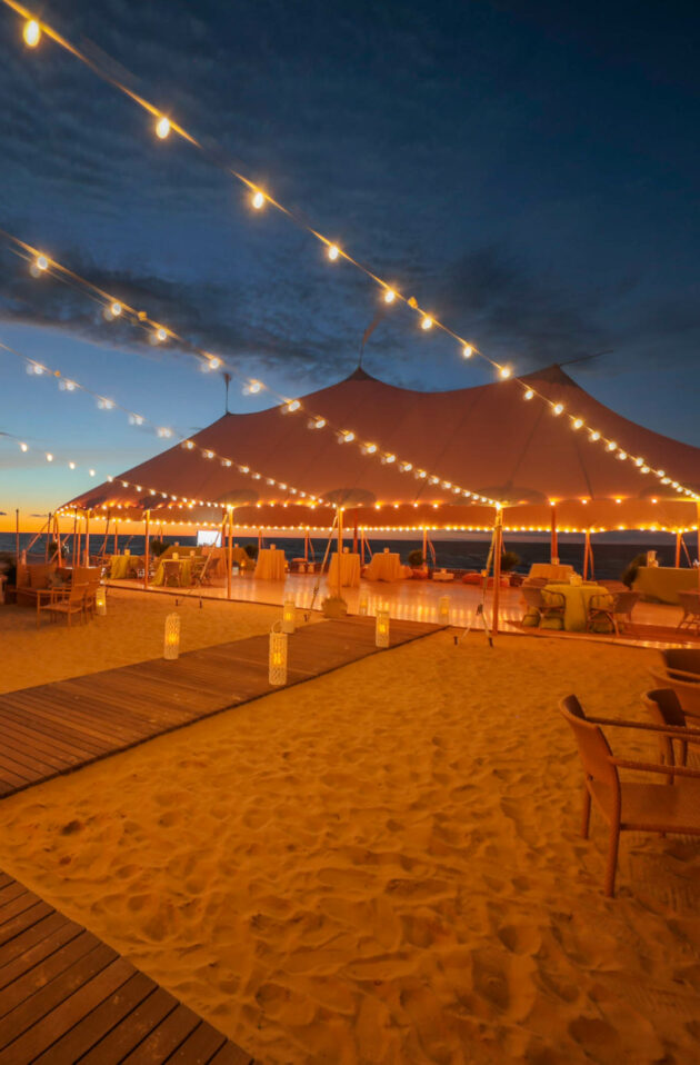 An outdoor evening event venue space lit by string lights under a large tent on a sandy beach at dusk.