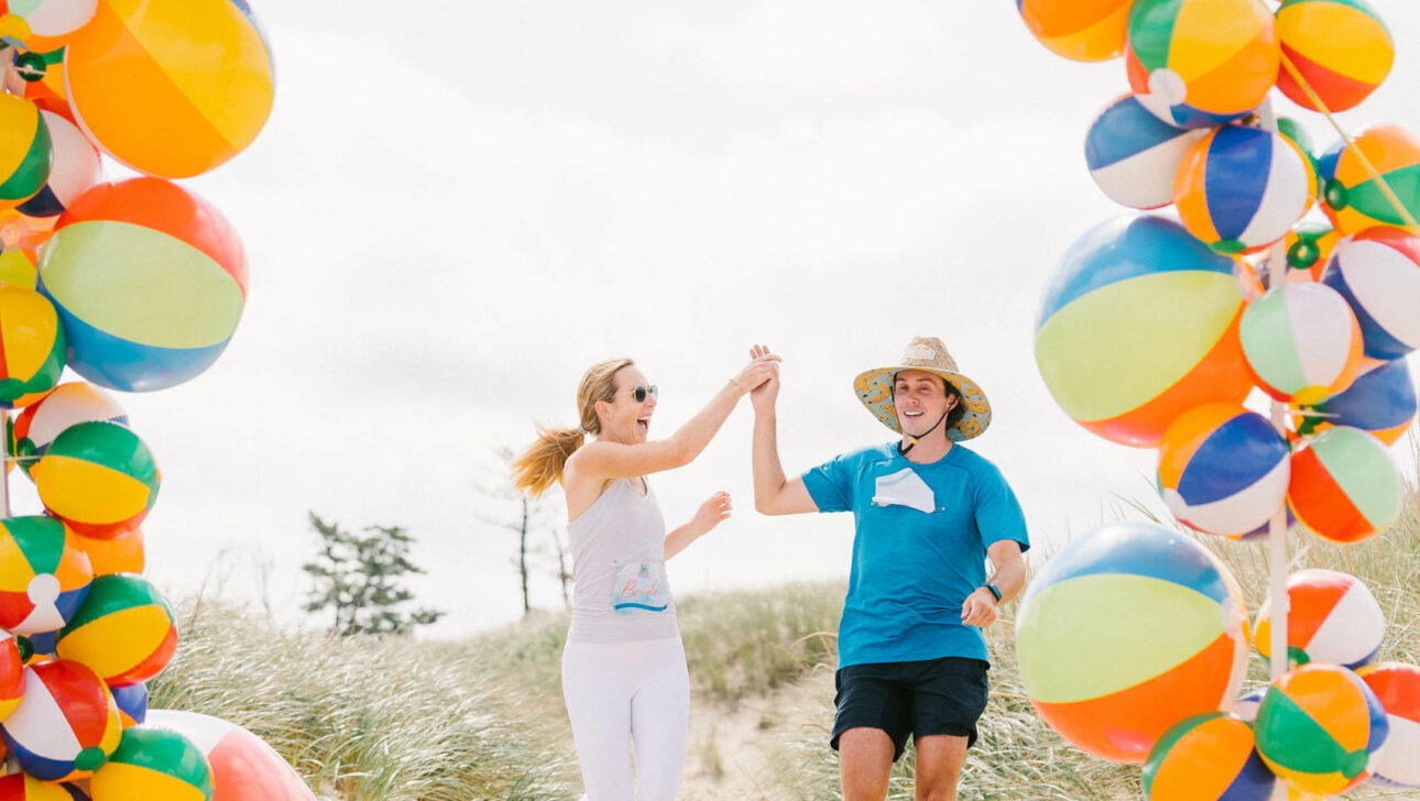 Two people joyfully running through a colorful beach ball arch on the sand at an event venue space.