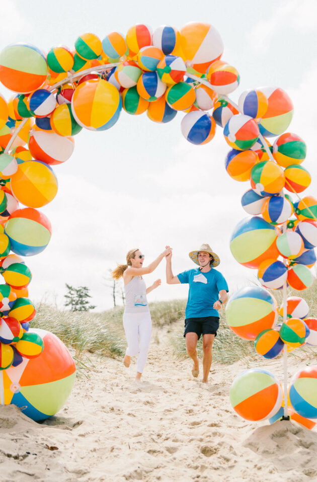 Two people joyfully running through a colorful beach ball arch on the sand at an event venue space.