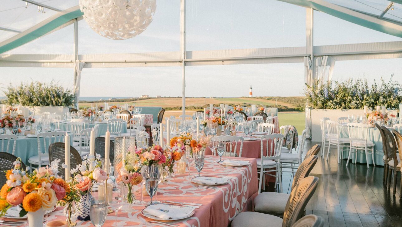 Elegant outdoor wedding reception setup with floral centerpieces under a tent overlooking a coastal landscape at dusk, meticulously organized by expert event planners.