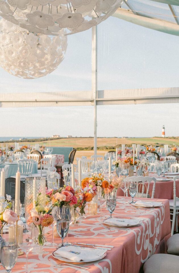 Elegant outdoor wedding reception setup with floral centerpieces under a tent overlooking a coastal landscape at dusk, meticulously organized by expert event planners.