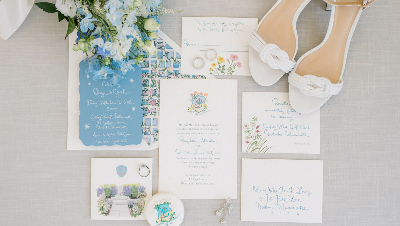 Wedding invitation suite with blue and floral motifs, matching accessories designed by an event planning company, and a pair of white bridal heels.