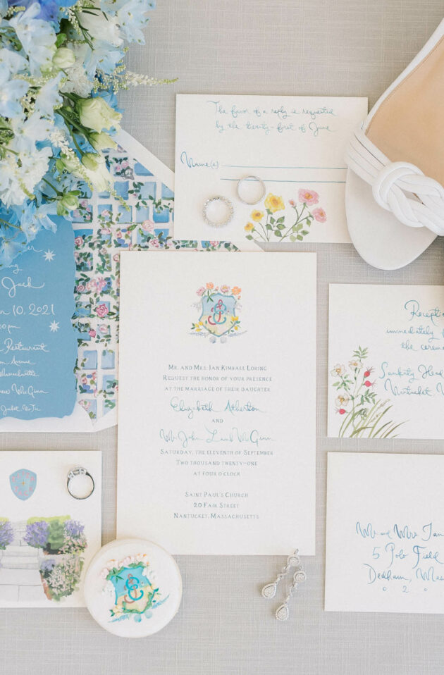 Wedding invitation suite with blue and floral motifs, matching accessories designed by an event planning company, and a pair of white bridal heels.