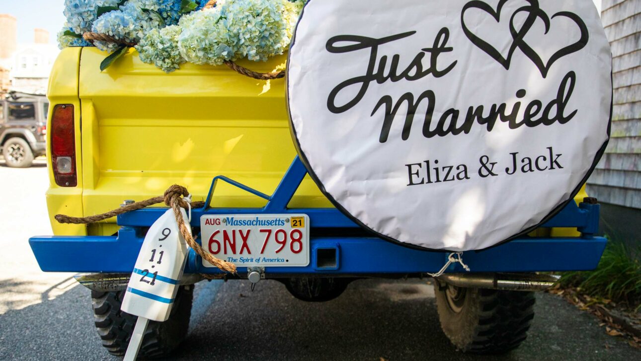 Yellow vehicle adorned with flowers and a "just married" sign featuring the names Eliza & Jack, with tin cans attached for a wedding celebration organized by an event planning company.