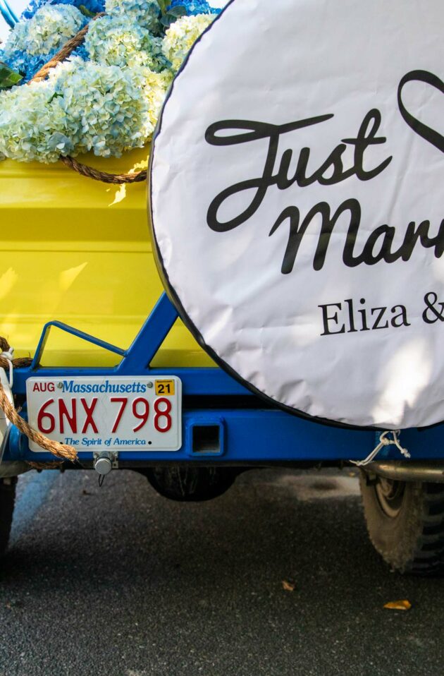 Yellow vehicle adorned with flowers and a "just married" sign featuring the names Eliza & Jack, with tin cans attached for a wedding celebration organized by an event planning company.