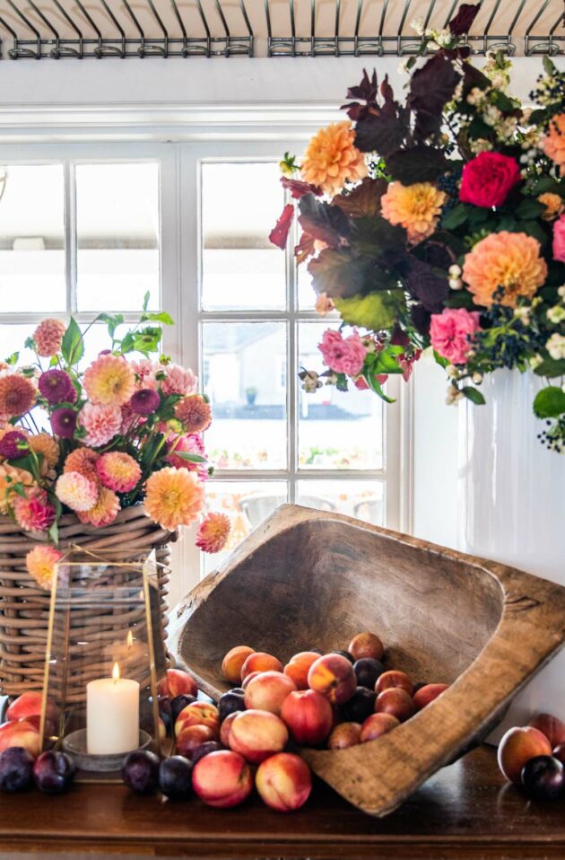 peaches rolling out of a wooden bowl on a table in front of a flower arrangement.
