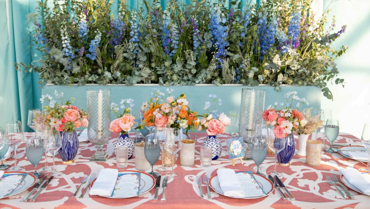 An elegantly set dining table at an event venue space with a floral centerpiece, blue and coral accents, and fine tableware.