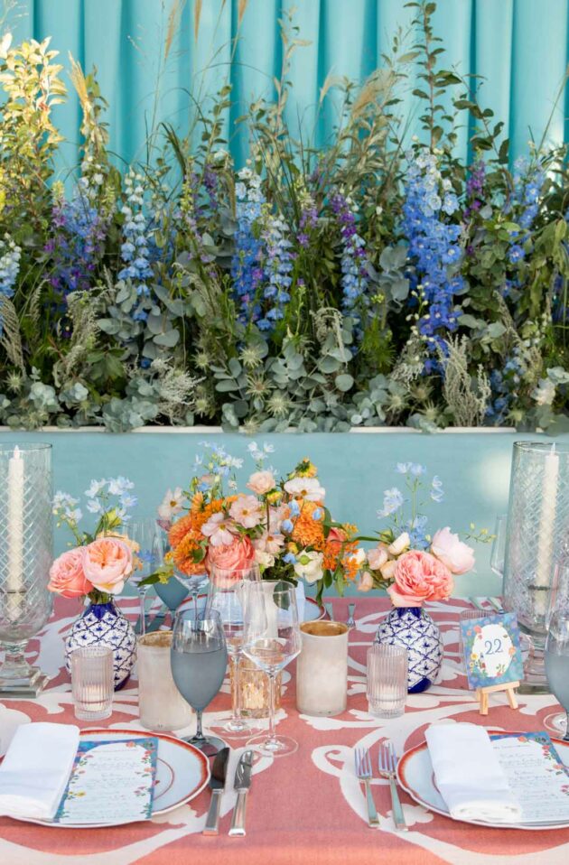 An elegantly set dining table at an event venue space with a floral centerpiece, blue and coral accents, and fine tableware.