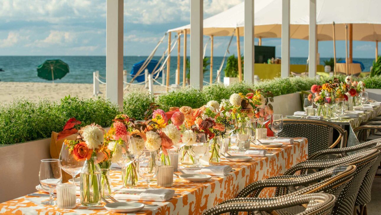 Beachfront dining setup with elegantly arranged tables, floral centerpieces, and a view of the ocean under a sunny sky, organized by a premier event planning company.