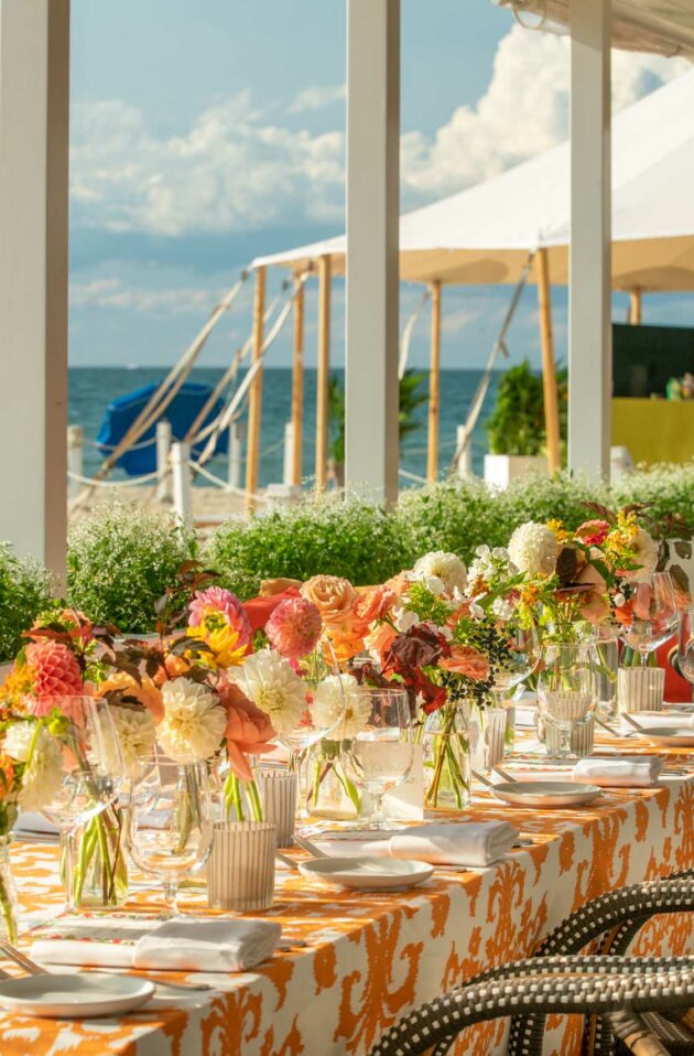 Beachfront dining setup with elegantly arranged tables, floral centerpieces, and a view of the ocean under a sunny sky, organized by a premier event planning company.