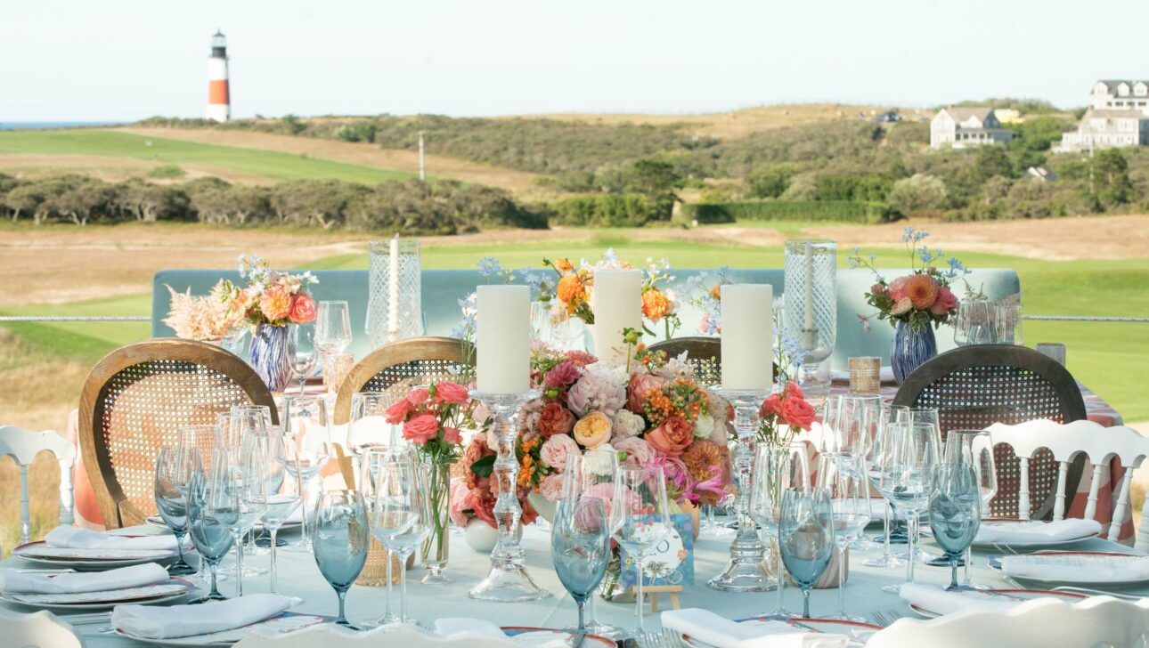 An elegantly set outdoor dining table with floral arrangements, organized by a premier event planning company, overlooking a coastal landscape with a lighthouse.