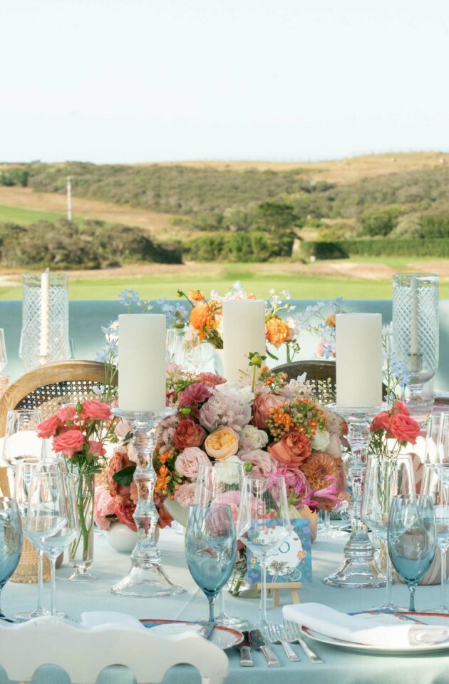 An elegantly set outdoor dining table with floral arrangements, organized by a premier event planning company, overlooking a coastal landscape with a lighthouse.