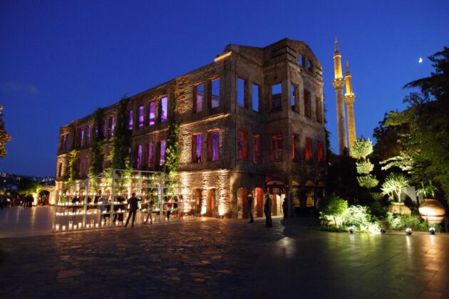 Historic event venue space illuminated at night with colorful lights, adjacent to a minaret under a twilight sky.