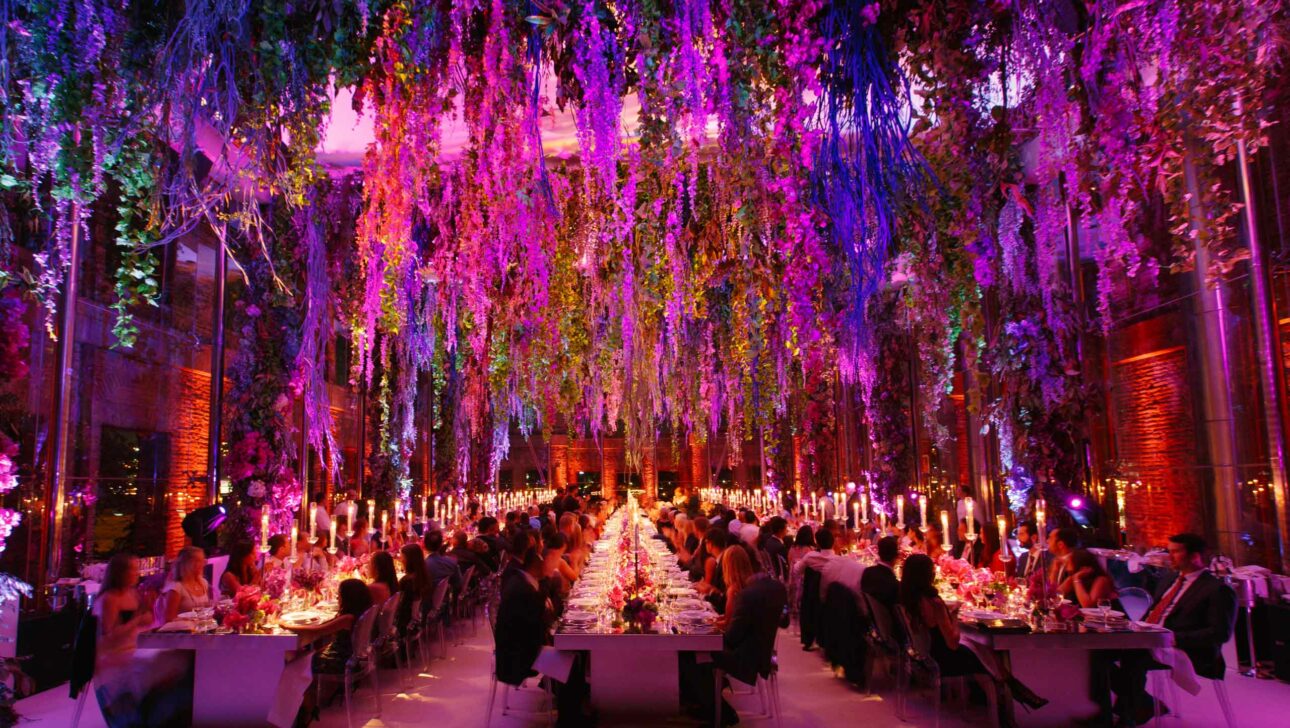 Elegant dinner event planned by a top event planning company, with guests seated at long tables amidst vibrant, colorful hanging floral decorations and atmospheric lighting.