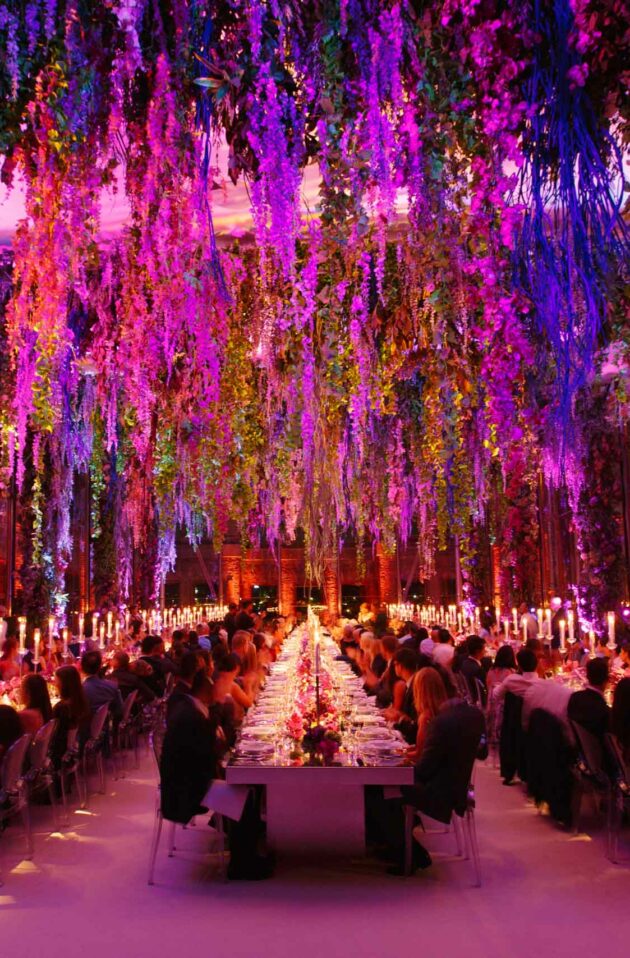 Elegant dinner event planned by a top event planning company, with guests seated at long tables amidst vibrant, colorful hanging floral decorations and atmospheric lighting.