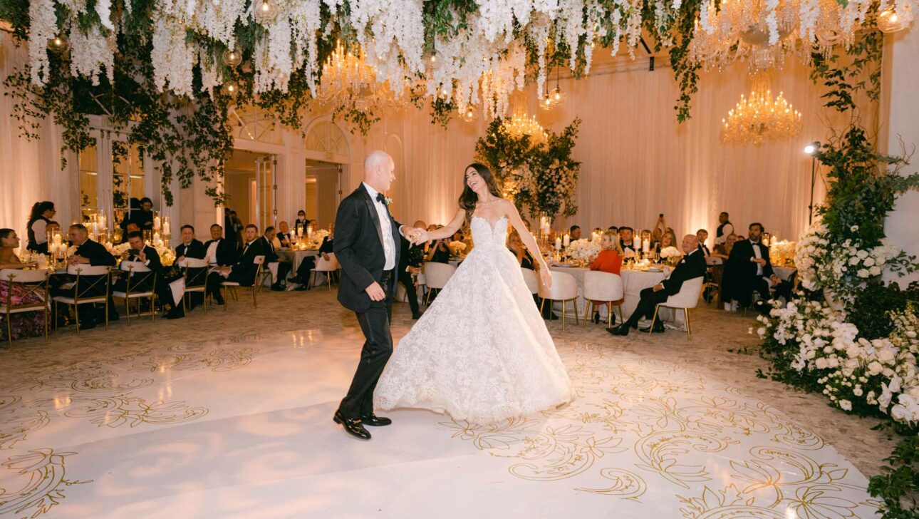 bride in wedding dress and man in tuxedo dancing in a reception hall full of people at tables.