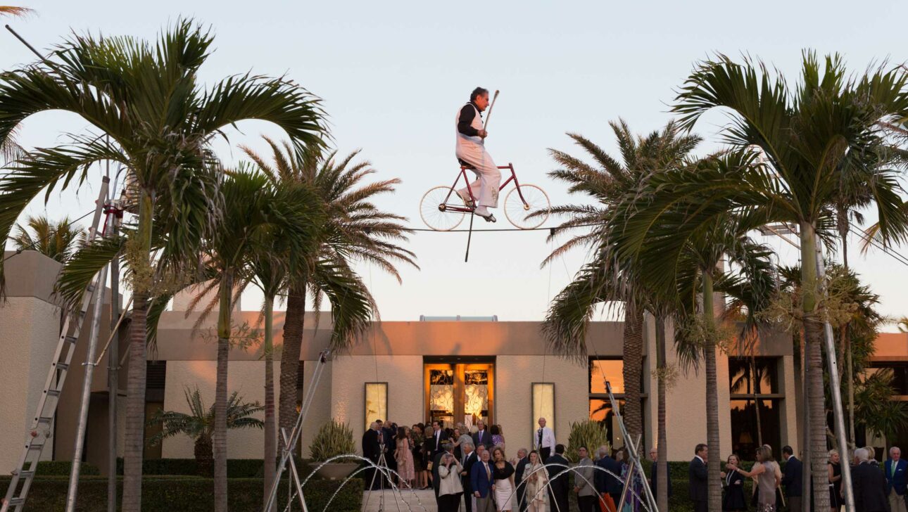 A performer riding a unicycle on a tightrope above a small fountain, with spectators gathered at an outdoor event planning company venue during twilight.