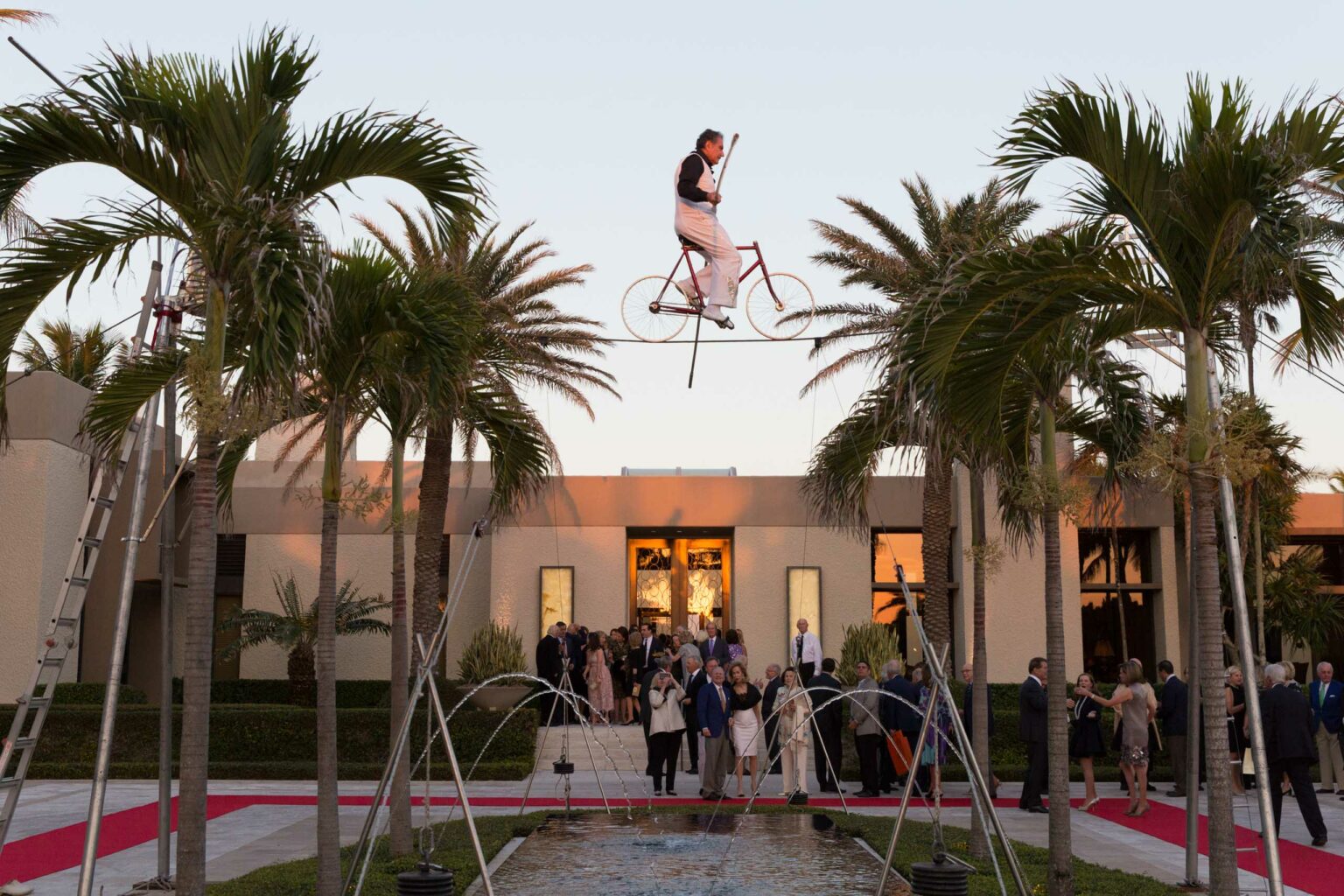 A performer riding a unicycle on a tightrope above a small fountain, with spectators gathered at an outdoor event planning company venue during twilight.