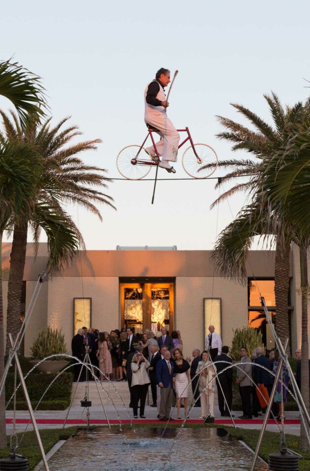 A performer riding a unicycle on a tightrope above a small fountain, with spectators gathered at an outdoor event planning company venue during twilight.