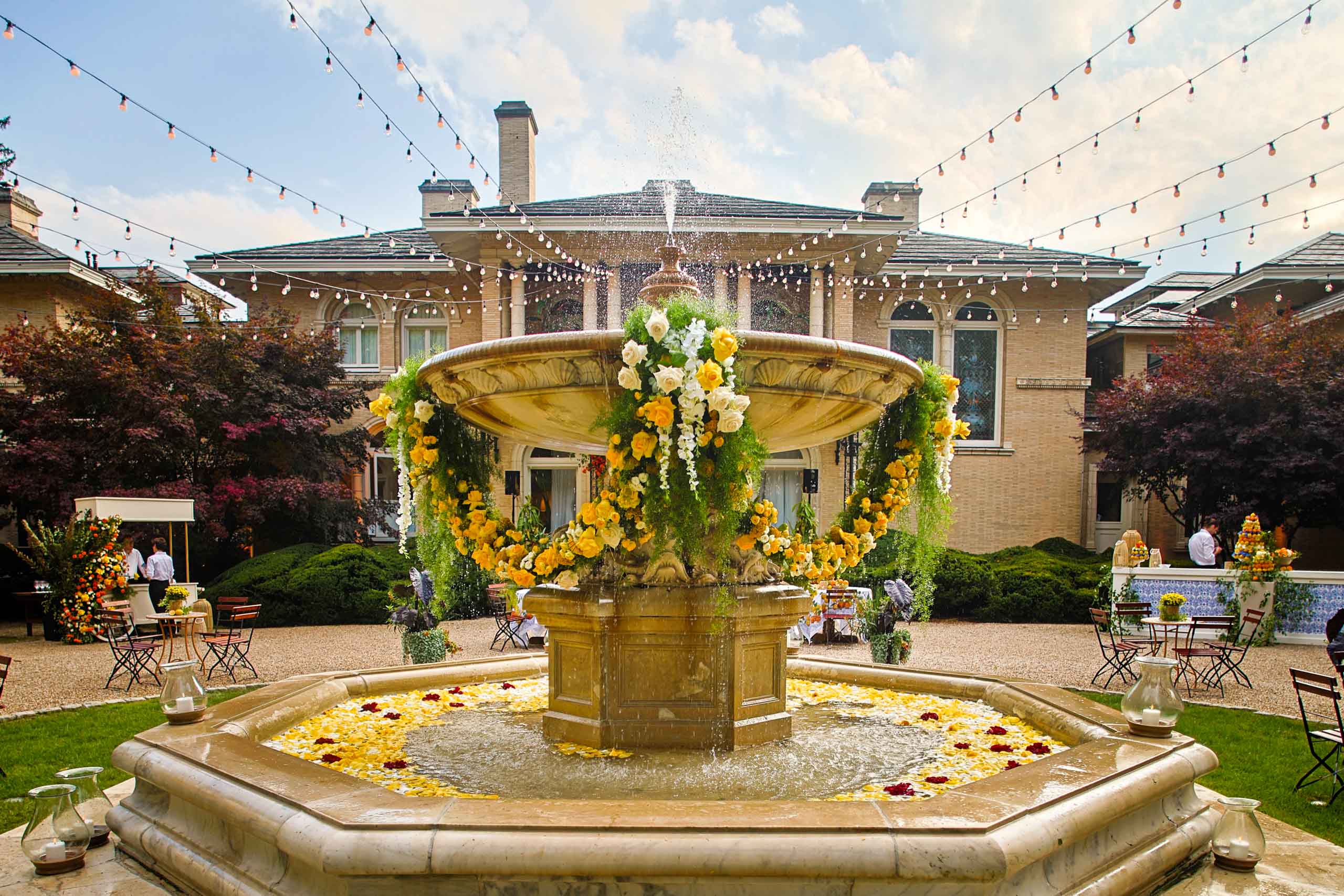 An ornate fountain adorned with yellow and white flowers, surrounded by an event venue space setting with string lights above.