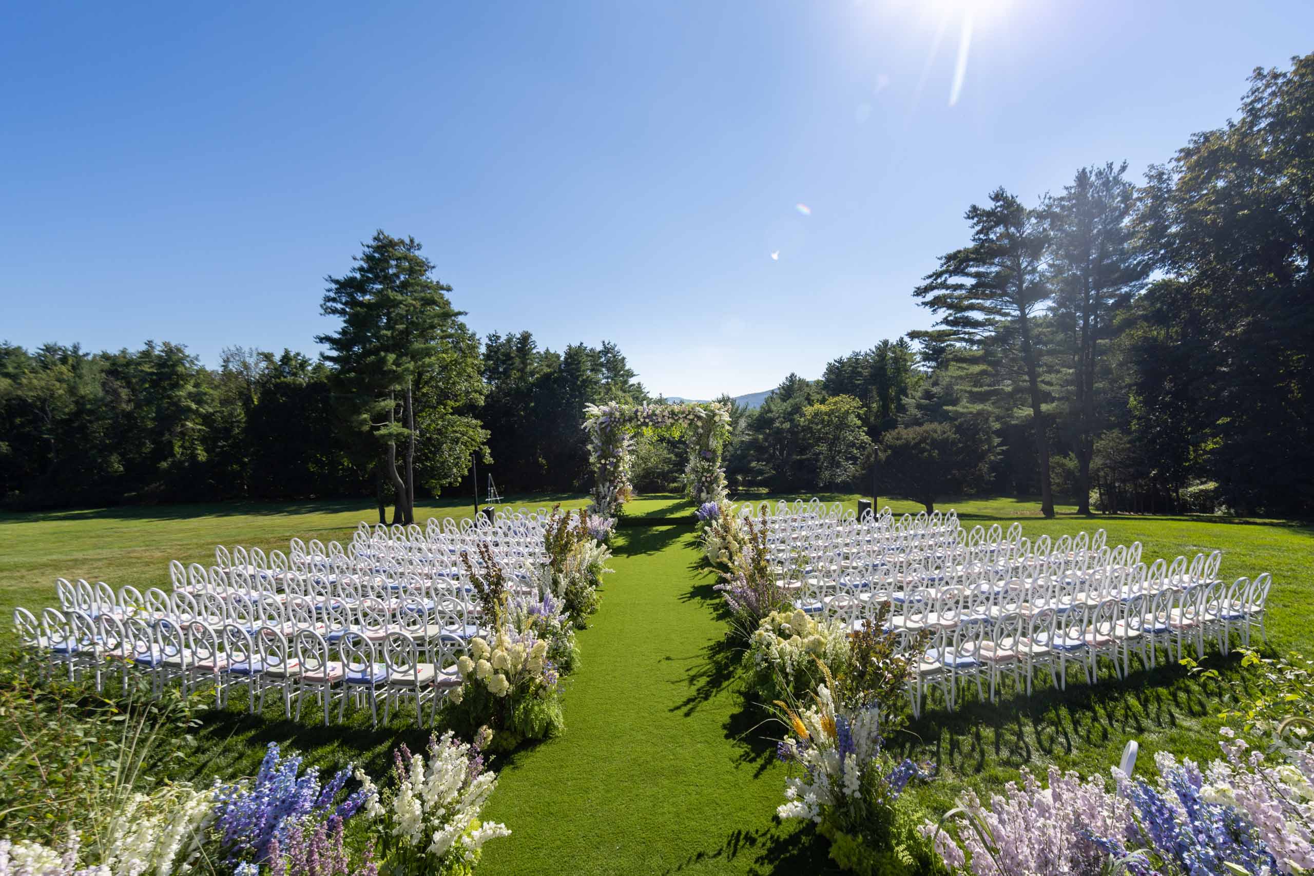 Outdoor wedding setup arranged by event planners with rows of white chairs and a floral aisle under clear skies.