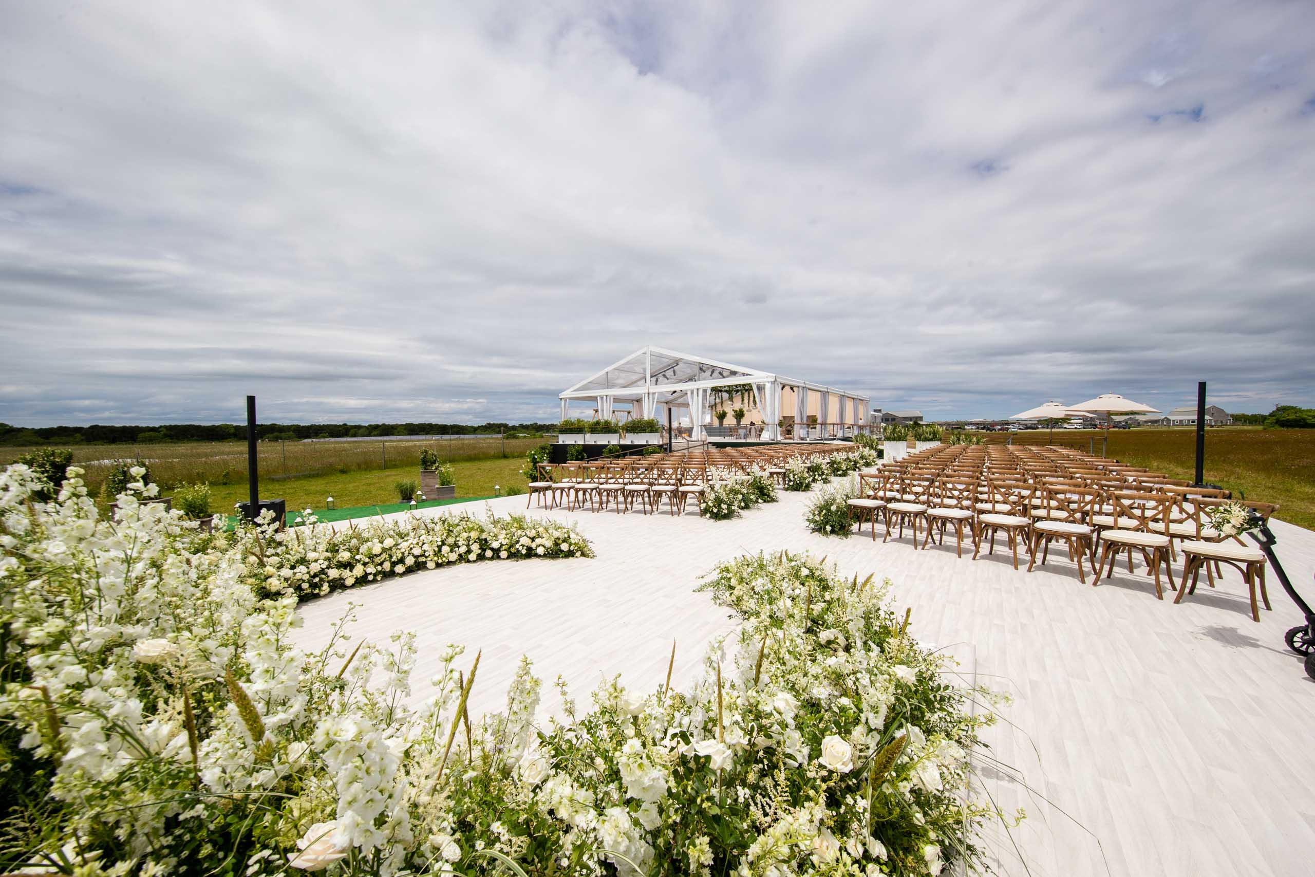 Outdoor wedding venue with white chairs and floral arrangements set up on a lawn under a cloudy sky, meticulously arranged by our top-notch event planning company.